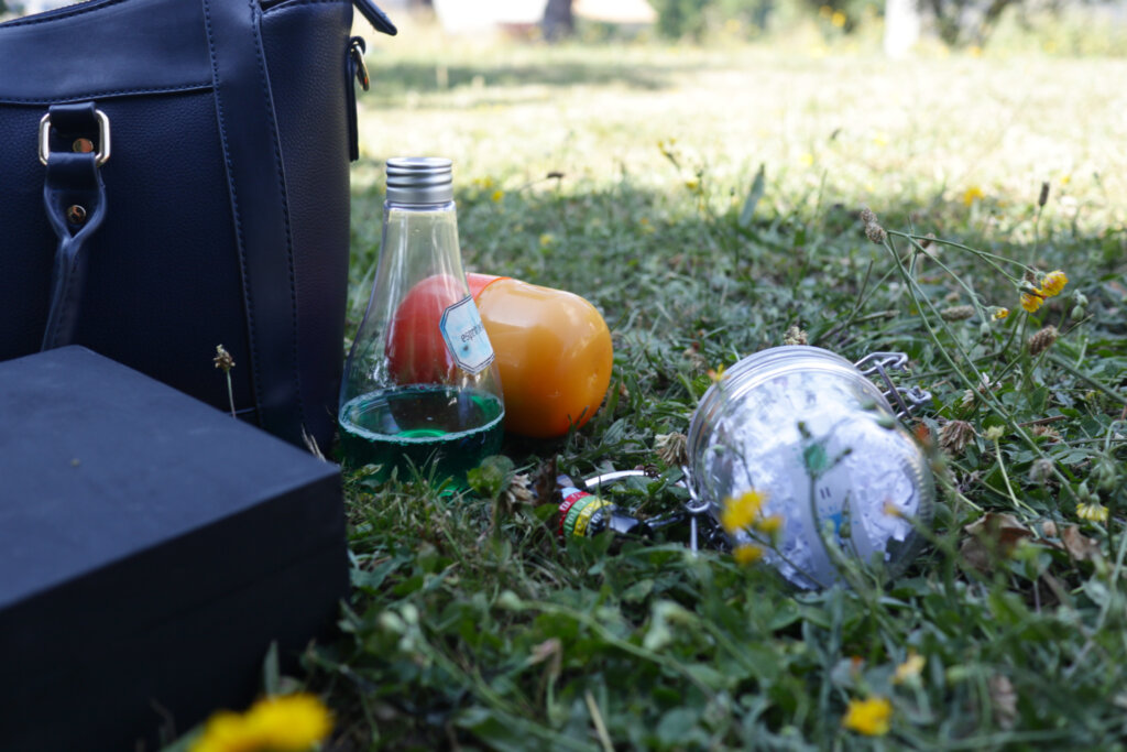 Photo de différents "indices" dans l'herbe lors d'une chasse au trésor / escape game à domicile. On peut voir une fiole contenant un liquide vert, un oeuf kinder orange, un cadenas à code, une boîte de bois fermée et un bocal de verre.